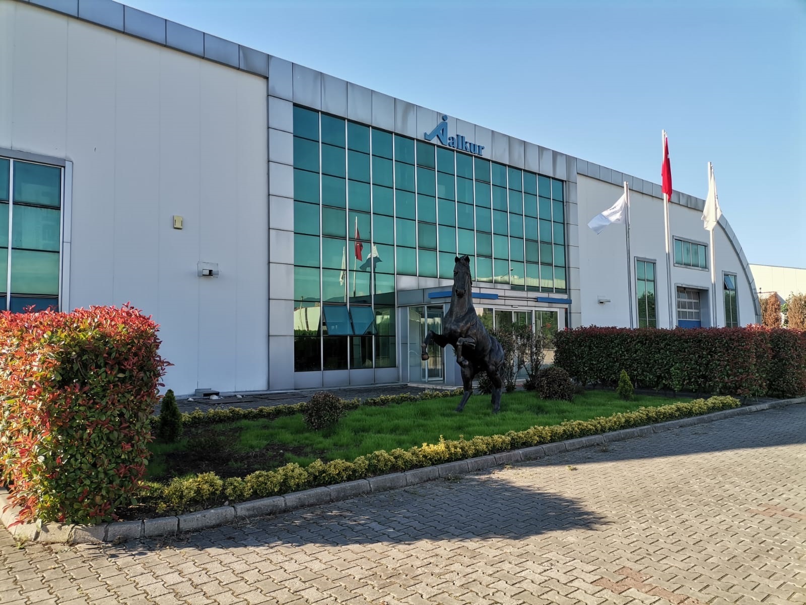 Alkur fabrika dış cephesi; cam cephe, logo ve giriş bahçesi.